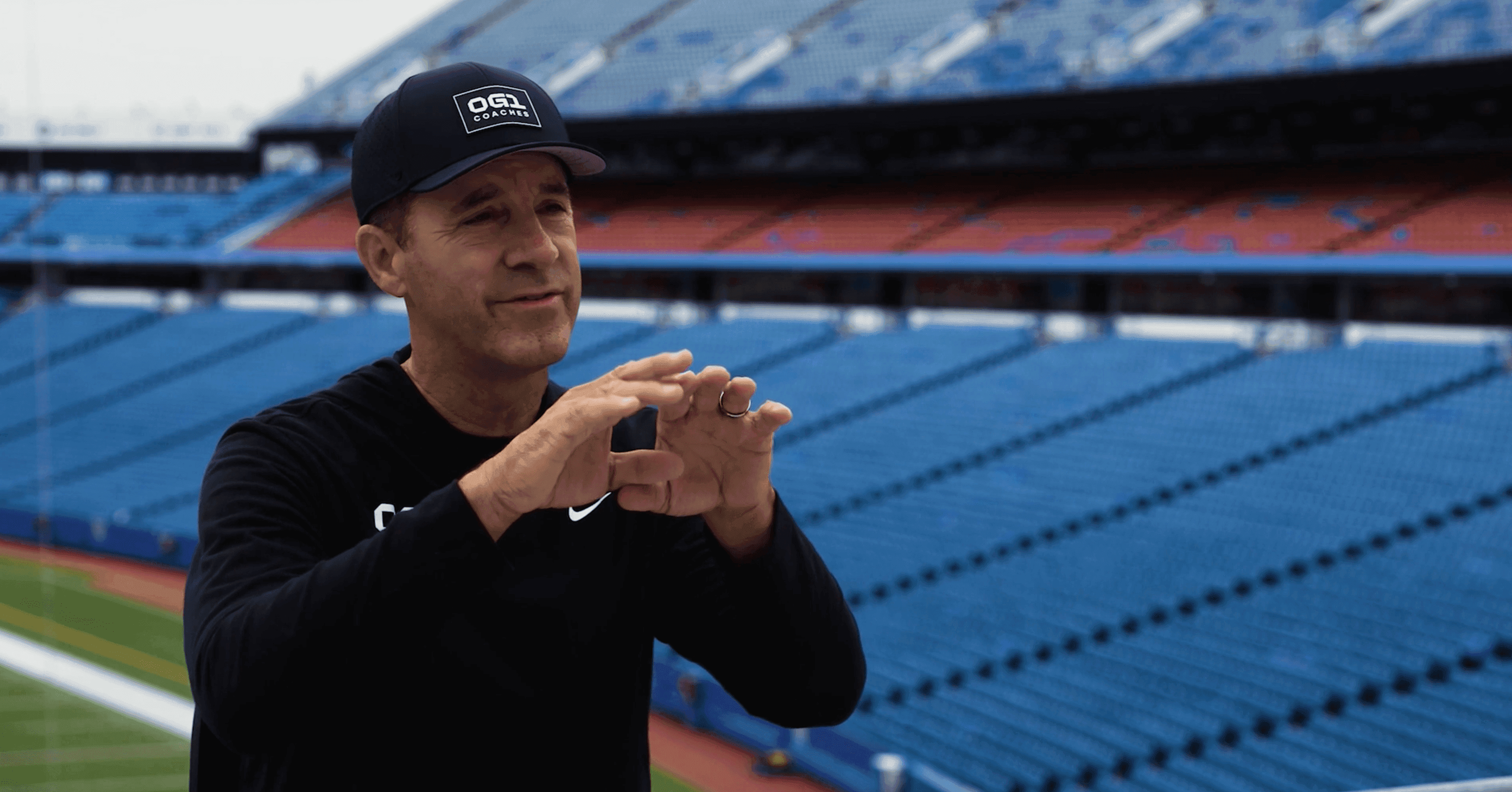 An OG1 coach in an OG1 cap on the sideline of an empty stadium.